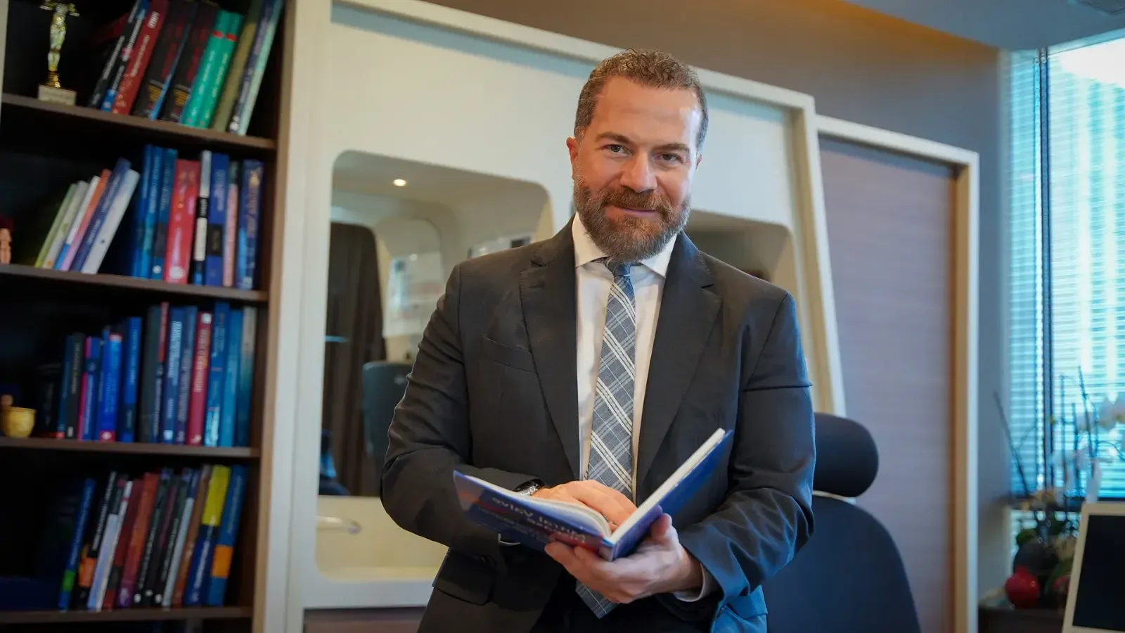 Prof. Dr. Ali Civelek kitaplığının önünde, elinde defter